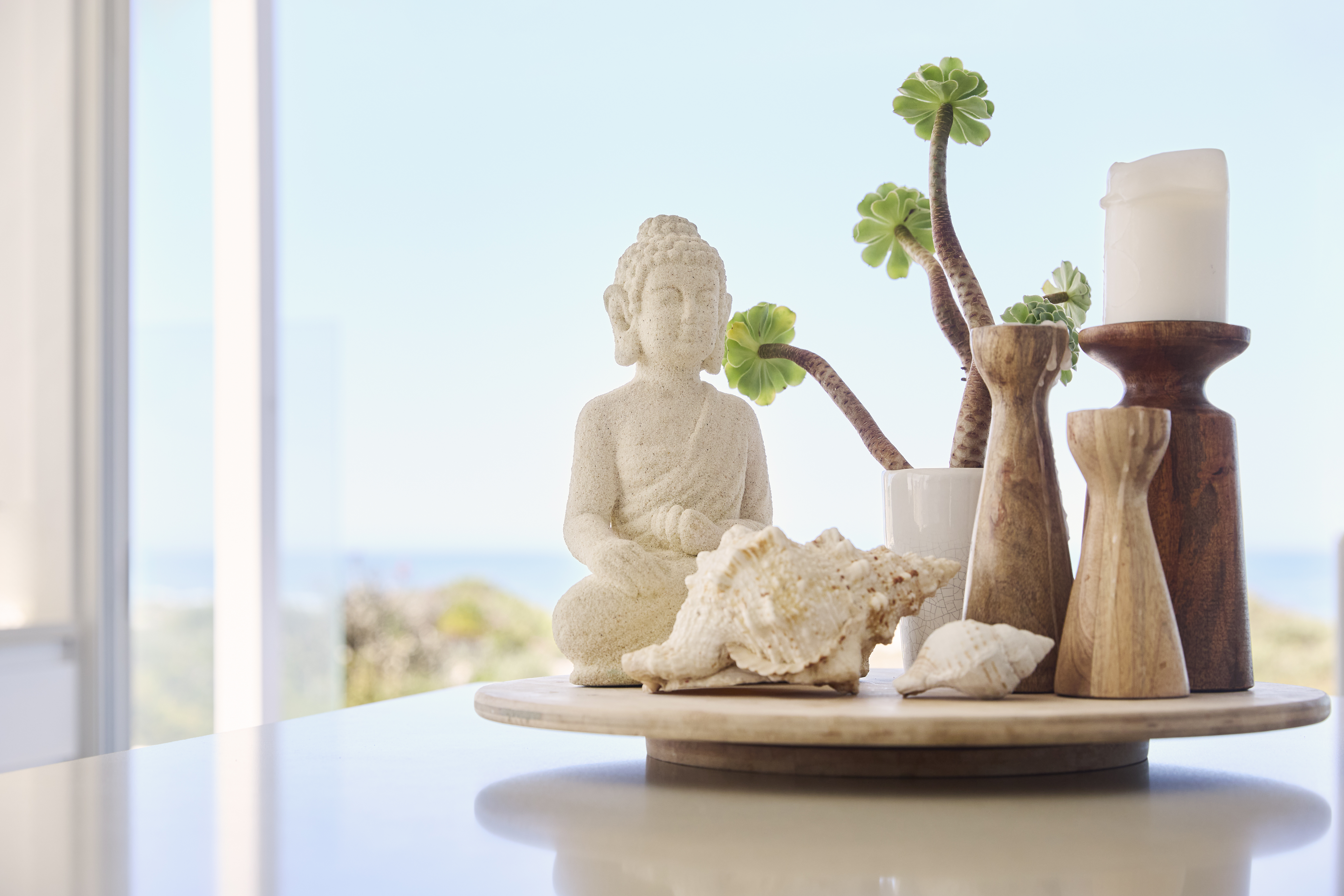 Decorative arrangement featuring a Buddha statue, seashells, wooden candle holders, and a succulent plant on a wooden tray with a beach view in the background.